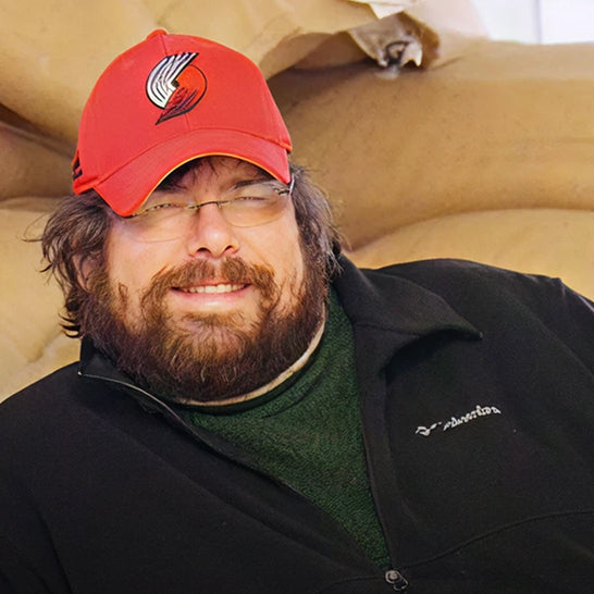 Man wearing a red cap with a logo and black jacket, sitting on a couch.