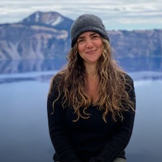 Woman with long hair and a beanie standing in front of a mountainous landscape.