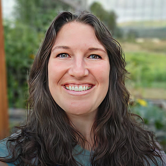 Woman with long brown hair smiling outdoors with greenery in the background