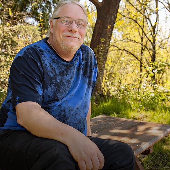 Man sitting outdoors on a wooden bench with trees and greenery in the background