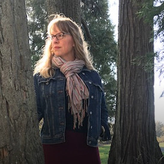 Woman wearing a denim jacket and plaid scarf standing between two trees.
