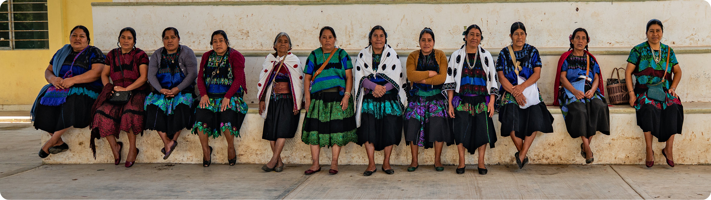 Row of women in traditional attire sitting against a building.